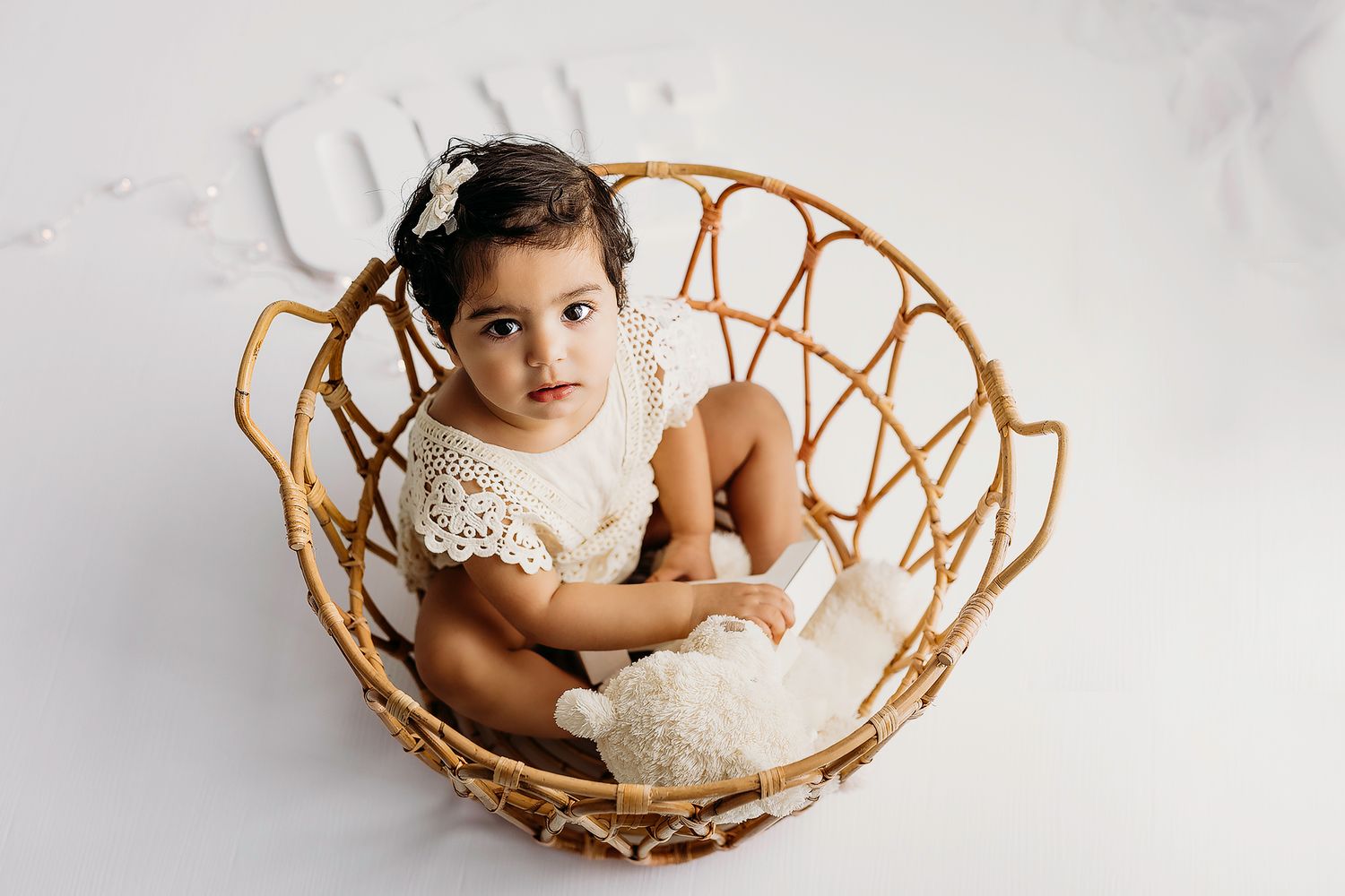 Small child in white lace dress sitting in round rattan peacock chair against white backdrop.
Rashinphotography-Nova photographer- DMV DC