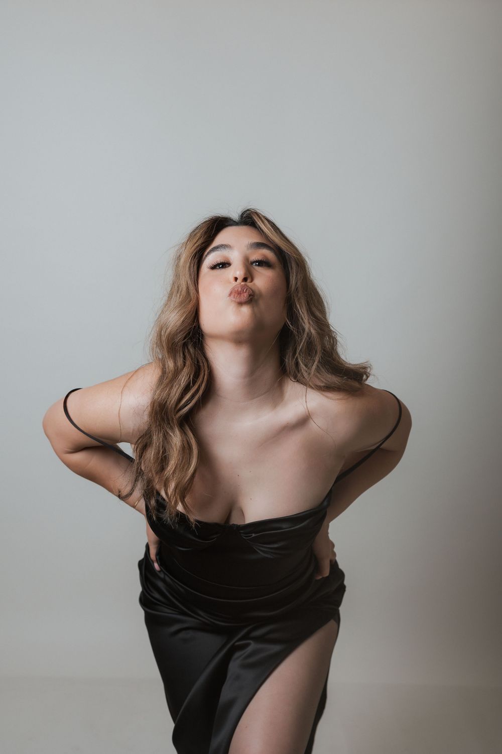 Playful pose in black evening dress with flowing hair against light backdrop.