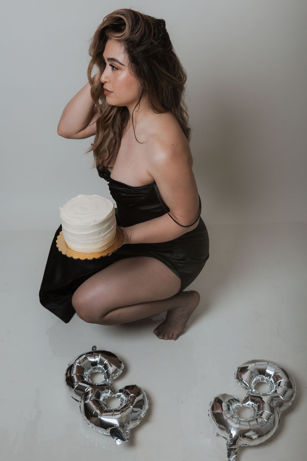 Side view of woman holding white cake while kneeling beside silver decorative pieces.