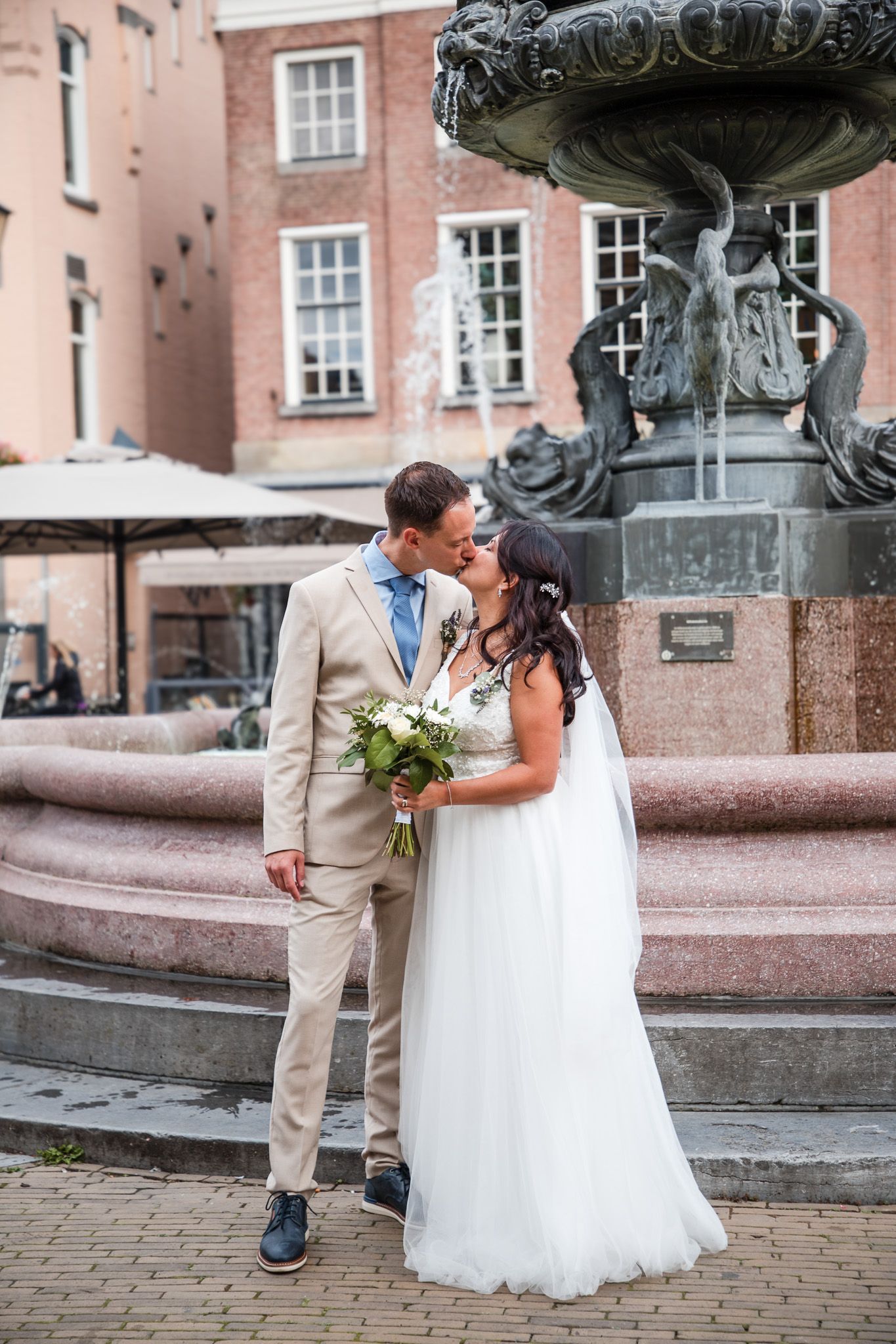 Newlyweds share romantic moments near an ornate stone fountain in front of a brick building after Gorinchem city hall wedding.