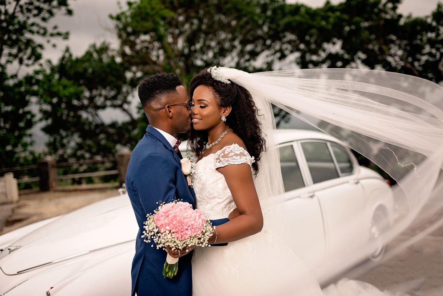 Romantic wedding portrait with flowing veil against white vintage car backdrop.