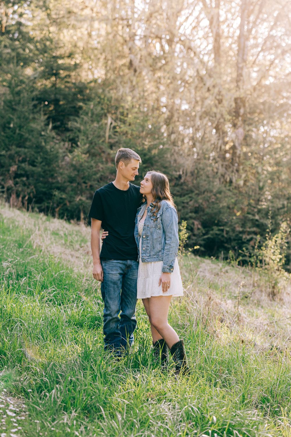 A couple stands together on a grassy hillside wearing casual attire on a sunny afternoon.