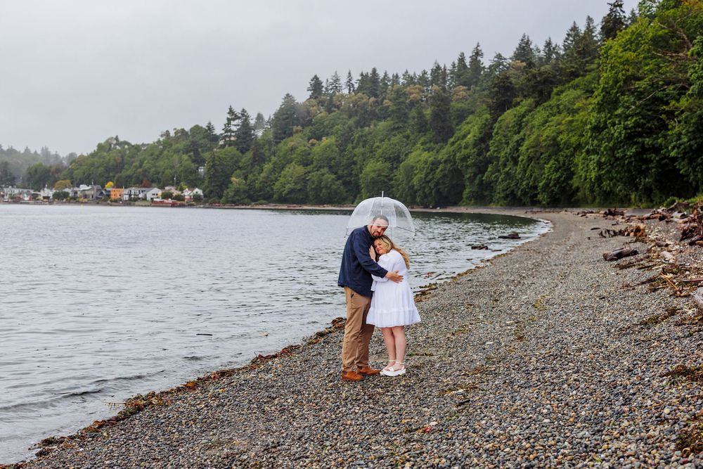 Two people embrace on a rocky beach shoreline with evergreen trees and houses along the water in the Pacific Northwest.