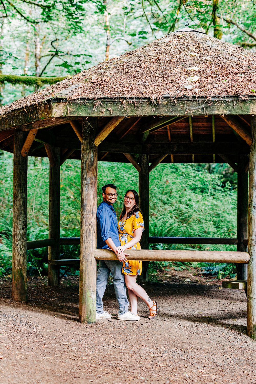 A rustic wooden gazebo with a brown shingled roof surrounded by lush green forest on a nature trail.