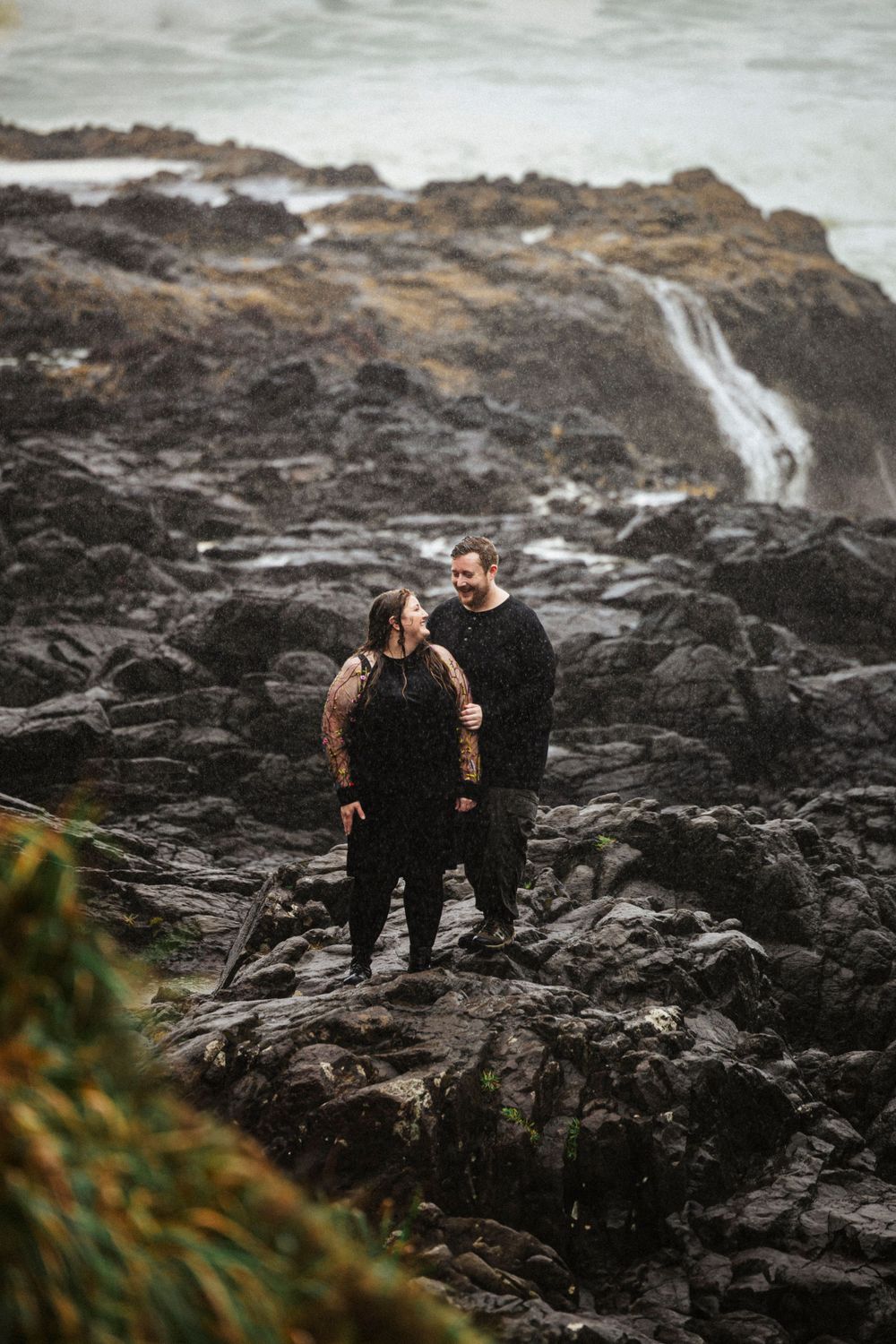 Two figures in dark clothing stand on rocky coastal cliffs with a misty waterfall cascading behind them.