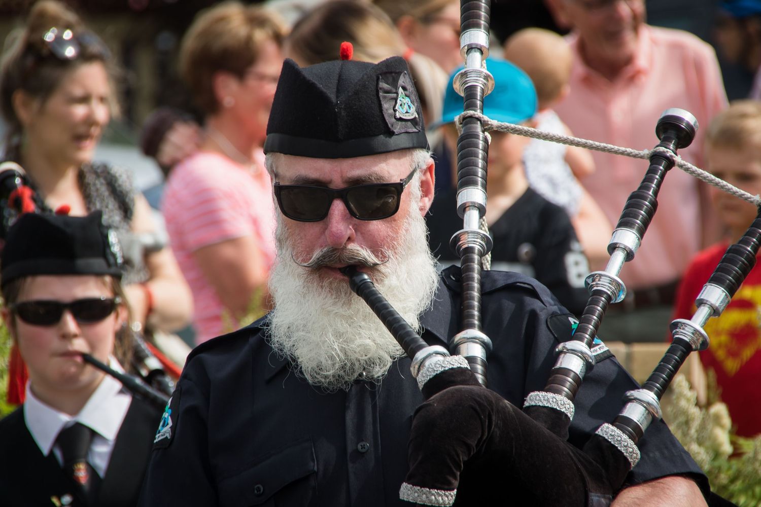 Bearded man in black uniform playing bagpipes at an outdoor parade event.