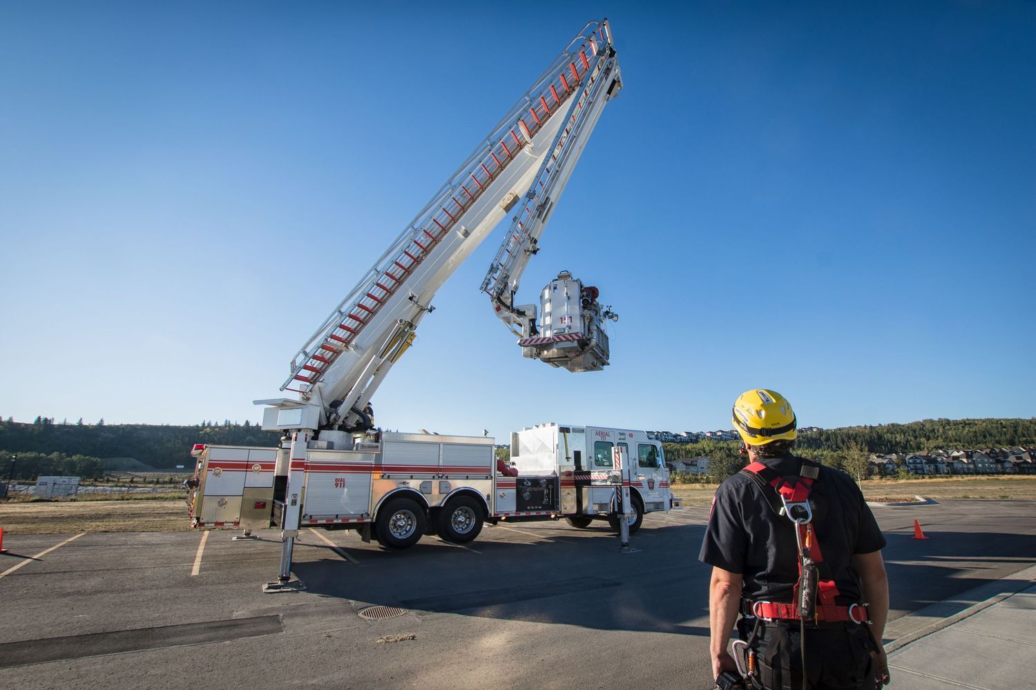 Firefighter in safety harness observing aerial ladder truck extended at outdoor training exercise.