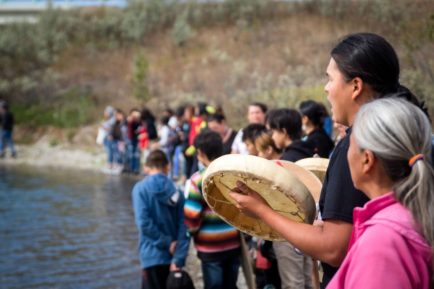Indigenous woman holding drum during traditional riverside ceremony with community gathering.