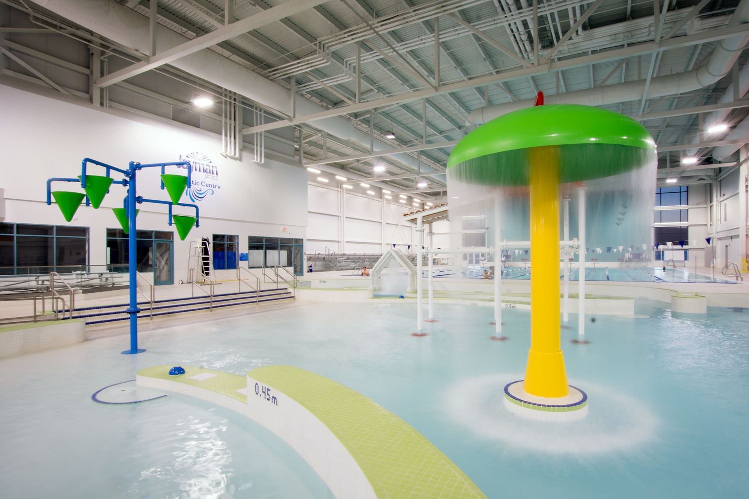 Indoor aquatic center featuring colorful splash pad with green mushroom water feature.