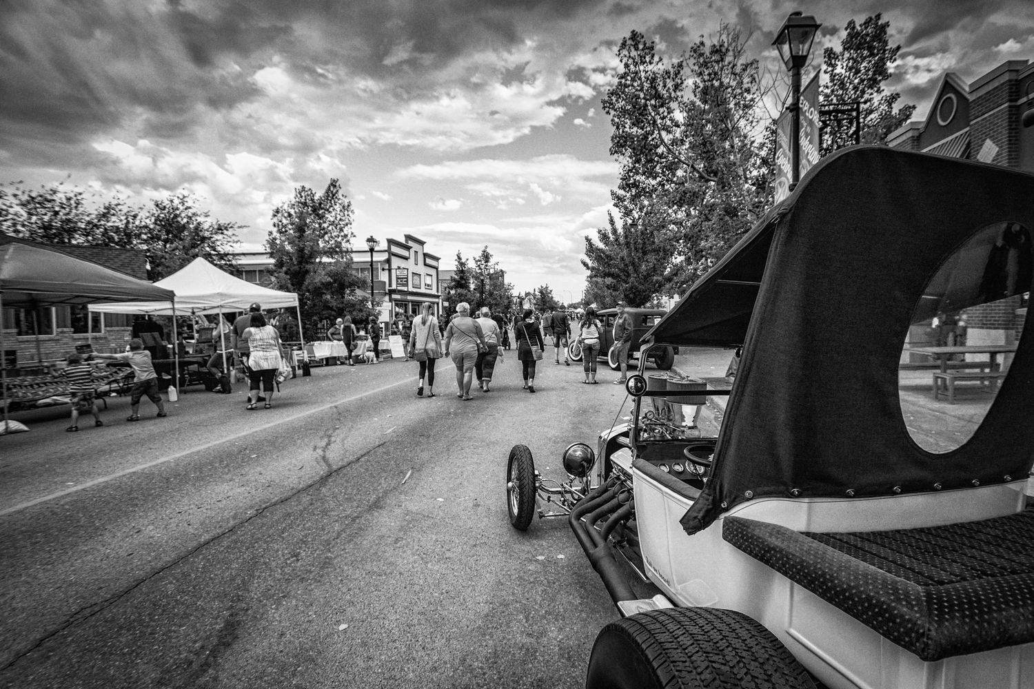Black and white street scene with vintage hot rod car at outdoor community festival.