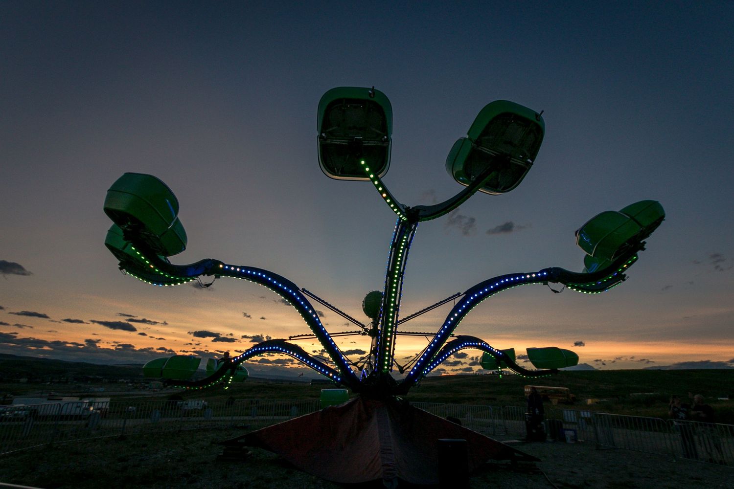Green illuminated carnival ride silhouetted against vibrant sunset sky at outdoor fair.