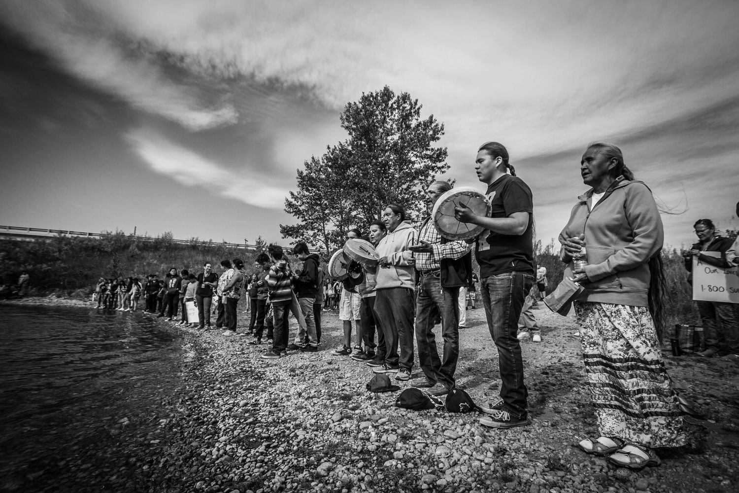 Black and white photo of Indigenous people drumming and singing in ceremony along a riverbank.