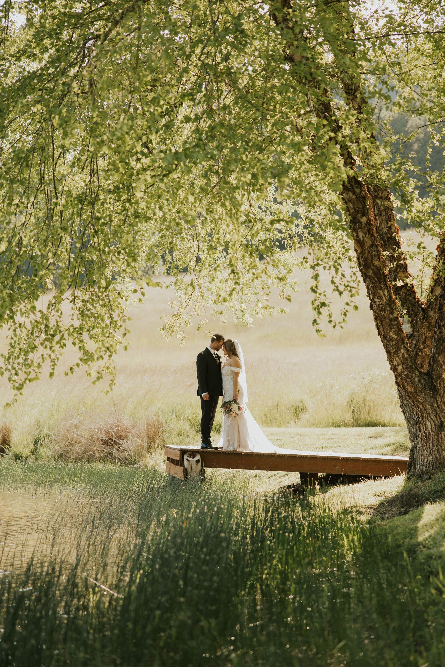 Bride and groom stand together under a large tree in a serene outdoor setting.