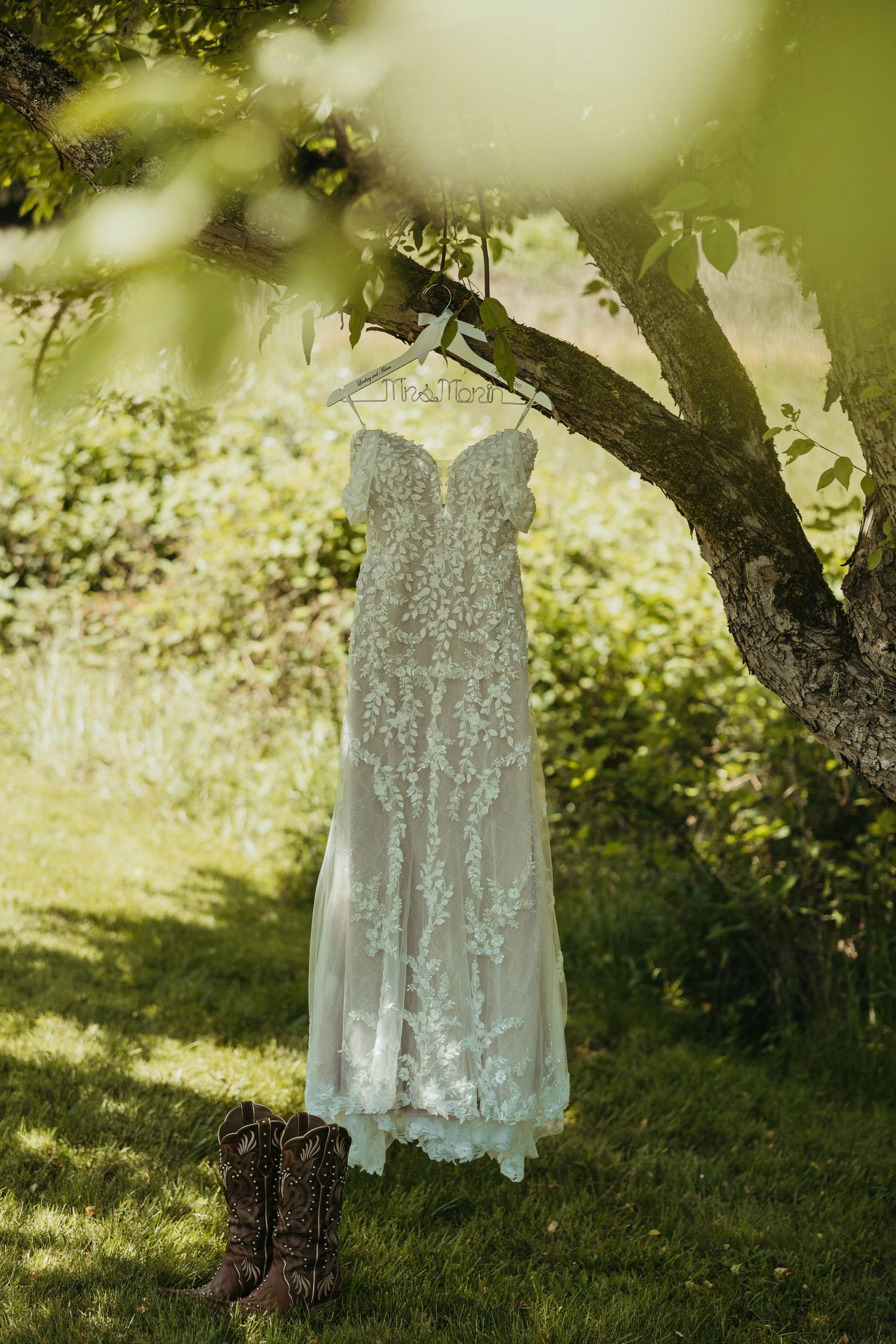 White lace wedding dress hangs ethereally under a tree in natural lighting.