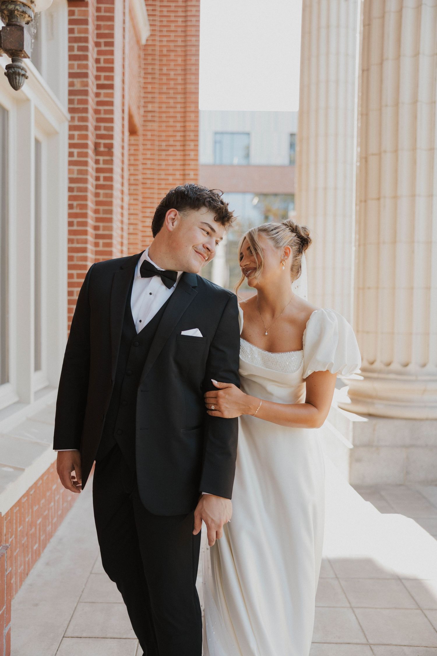Elegant couple in formal wedding attire poses playfully between classic brick columns.