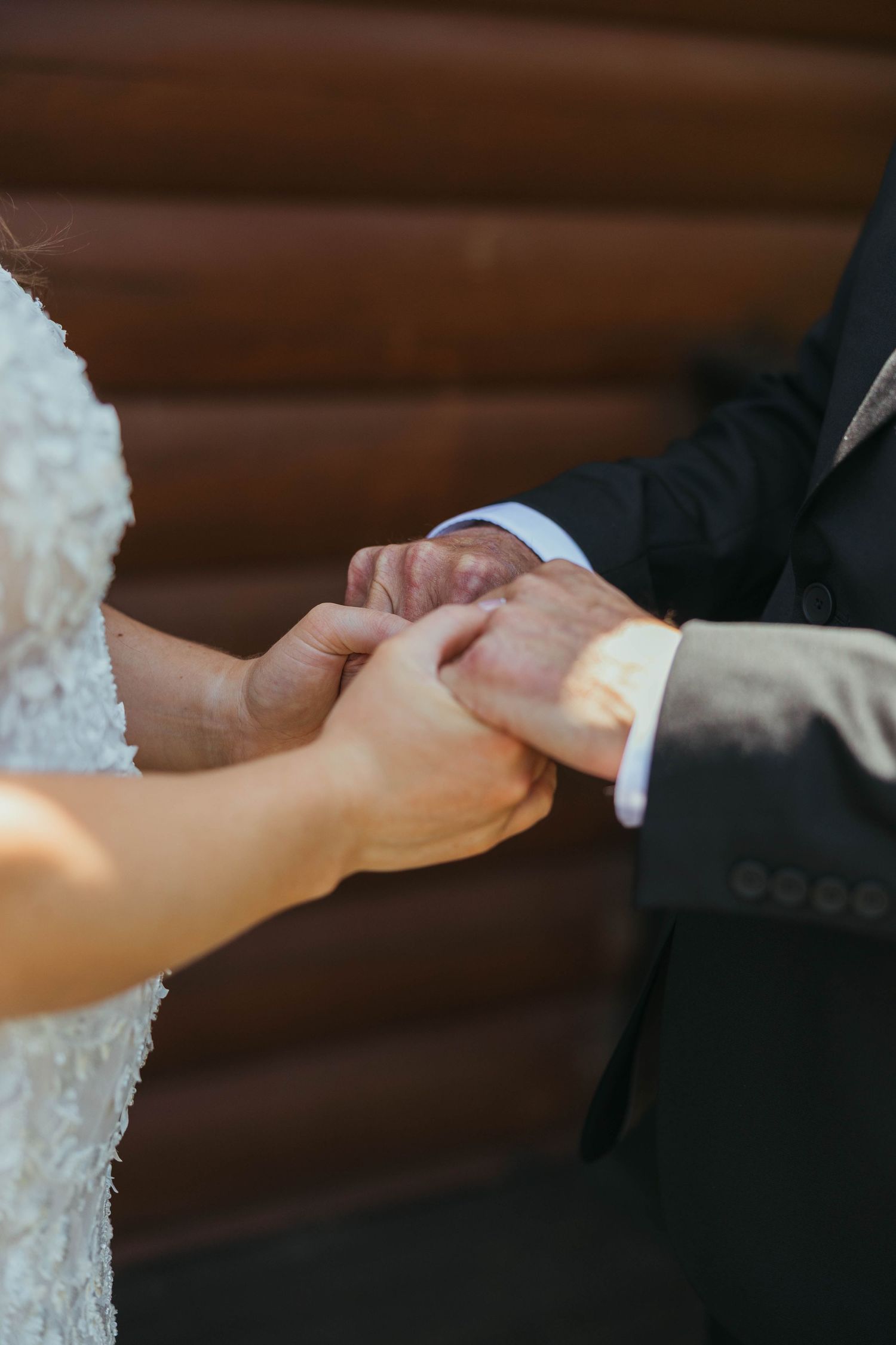 Close-up of bride and groom holding hands against wooden cabin wall.