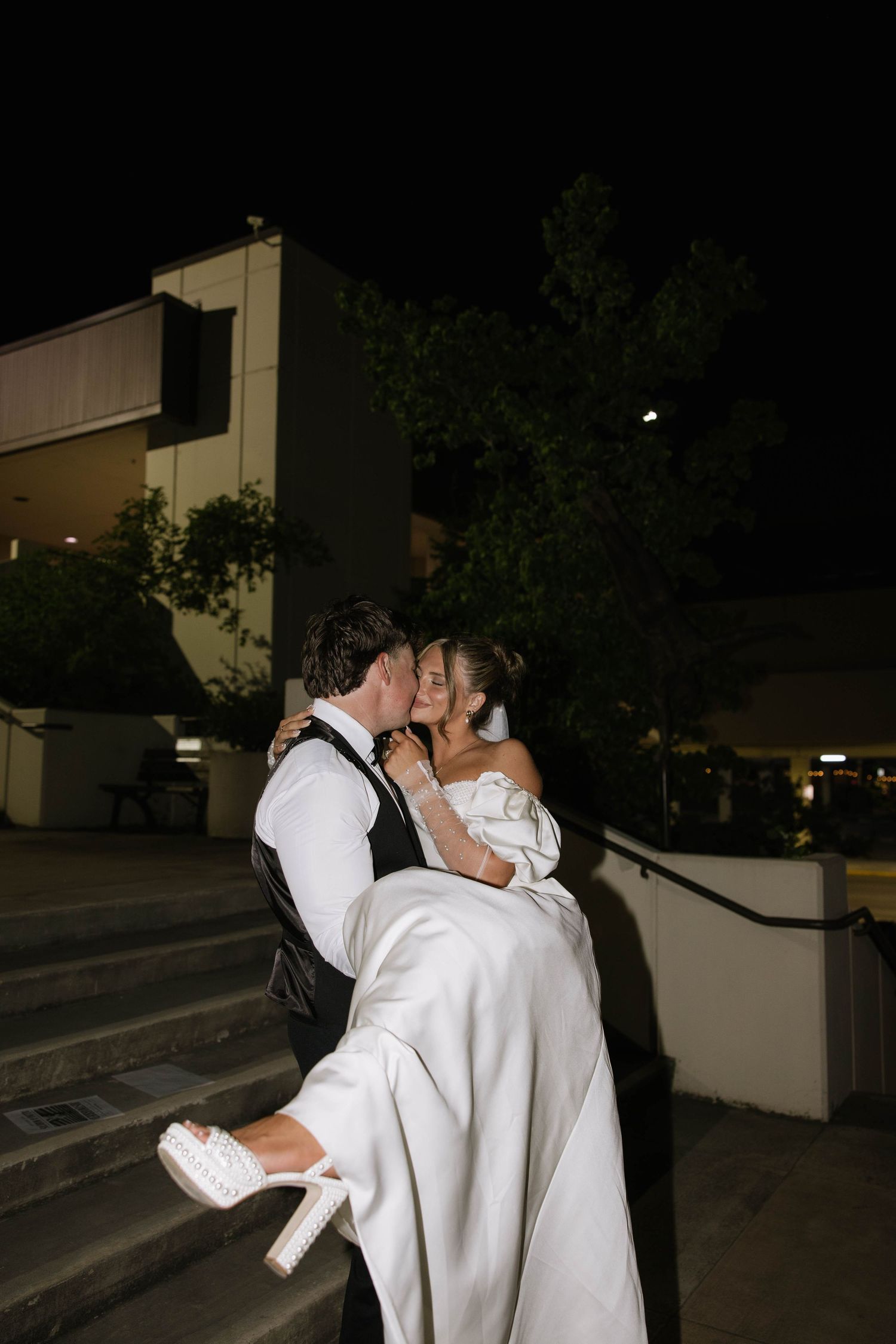 Romantic nighttime wedding couple embrace under outdoor lighting.