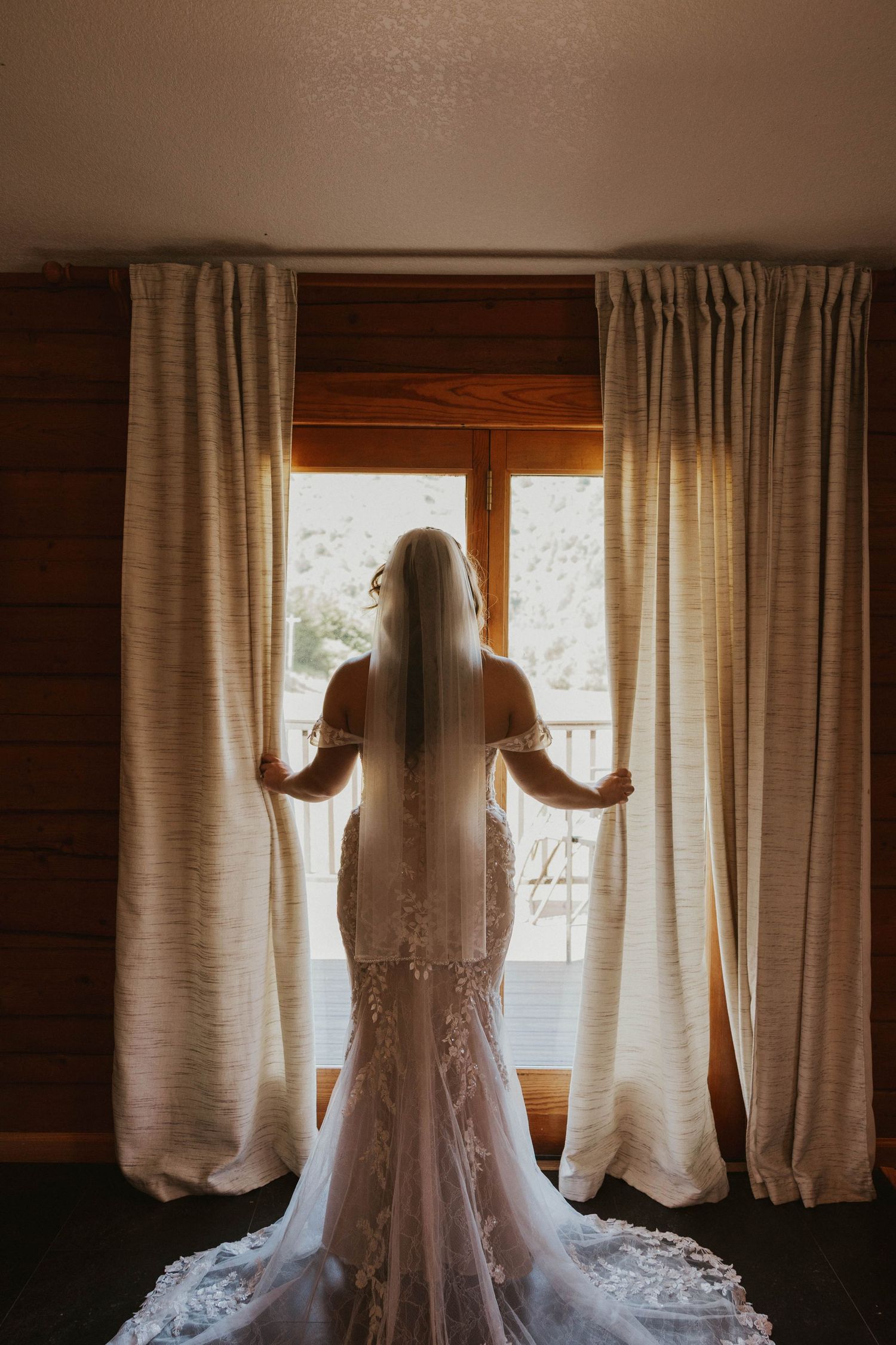 Silhouette of bride in lace gown standing by window with flowing curtains.