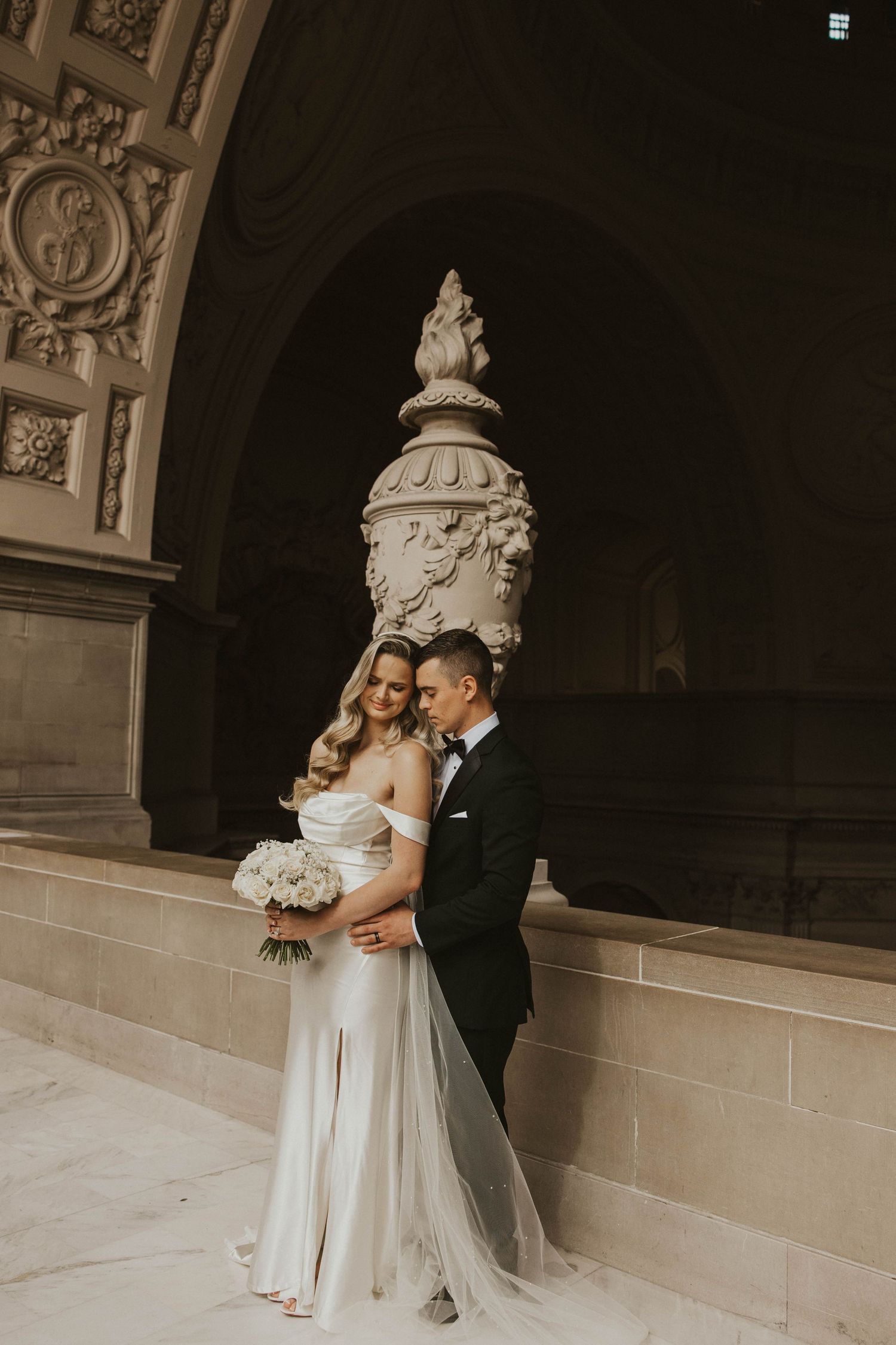 Wedding couple embracing in elegant stone hallway with ornate architectural details.