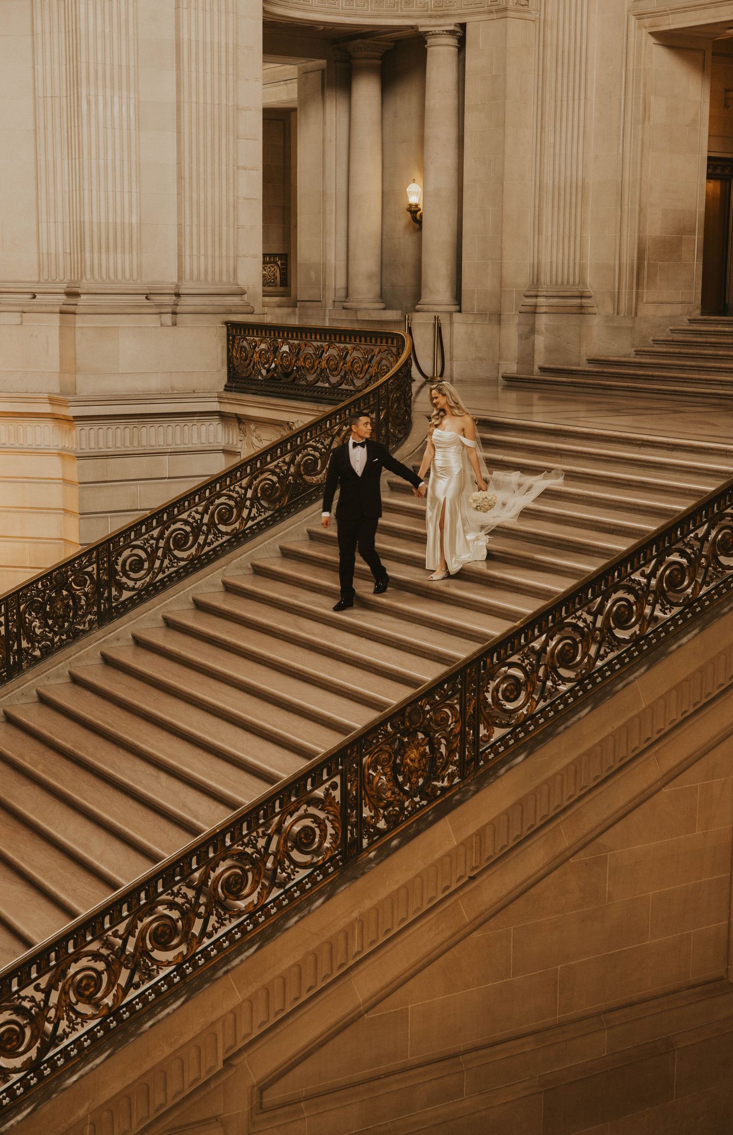 A couple descends an elegant grand staircase with ornate wrought iron railings in a classical building.