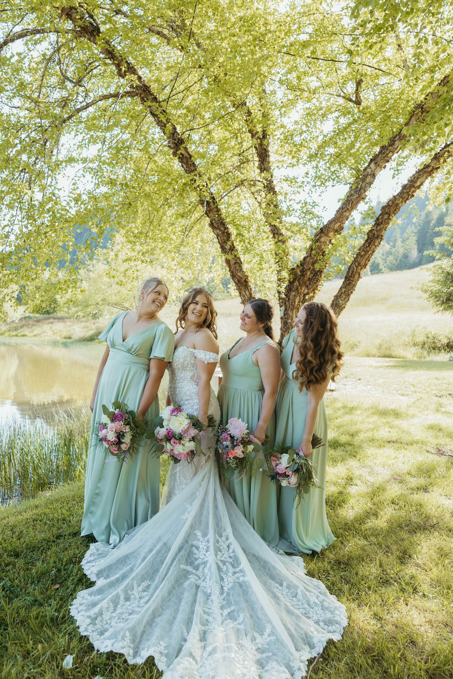 Wedding party in mint green dresses poses under blooming trees by a lake.