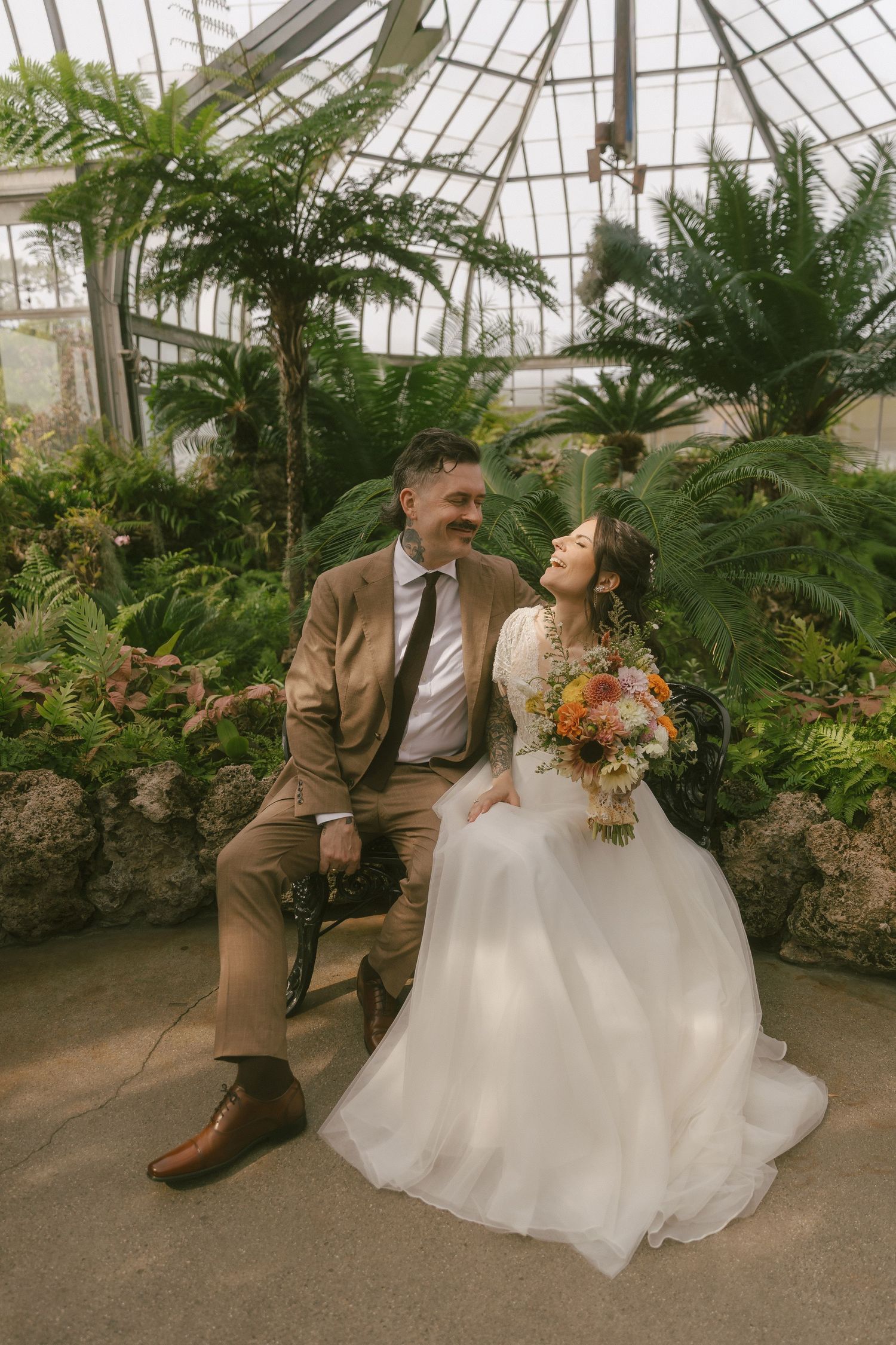 Two brides in flowing white gowns share a moment in front of a classic white farmhouse.