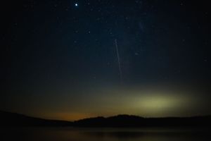 Meteor streaks across starlit sky over mountain silhouettes at dusk.