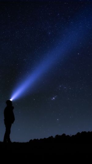 A silhouetted figure shines a bright blue flashlight beam into the starry night sky.