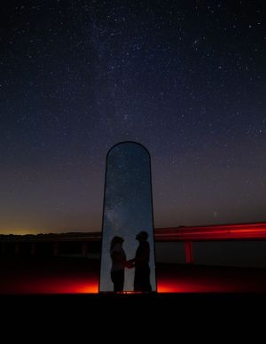 Silhouettes of two people standing by a tall silo under a starlit sky with red lighting.