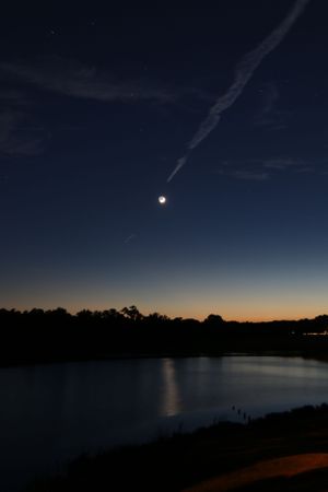 Moon setting over a lake at dusk with contrails in the darkening sky.