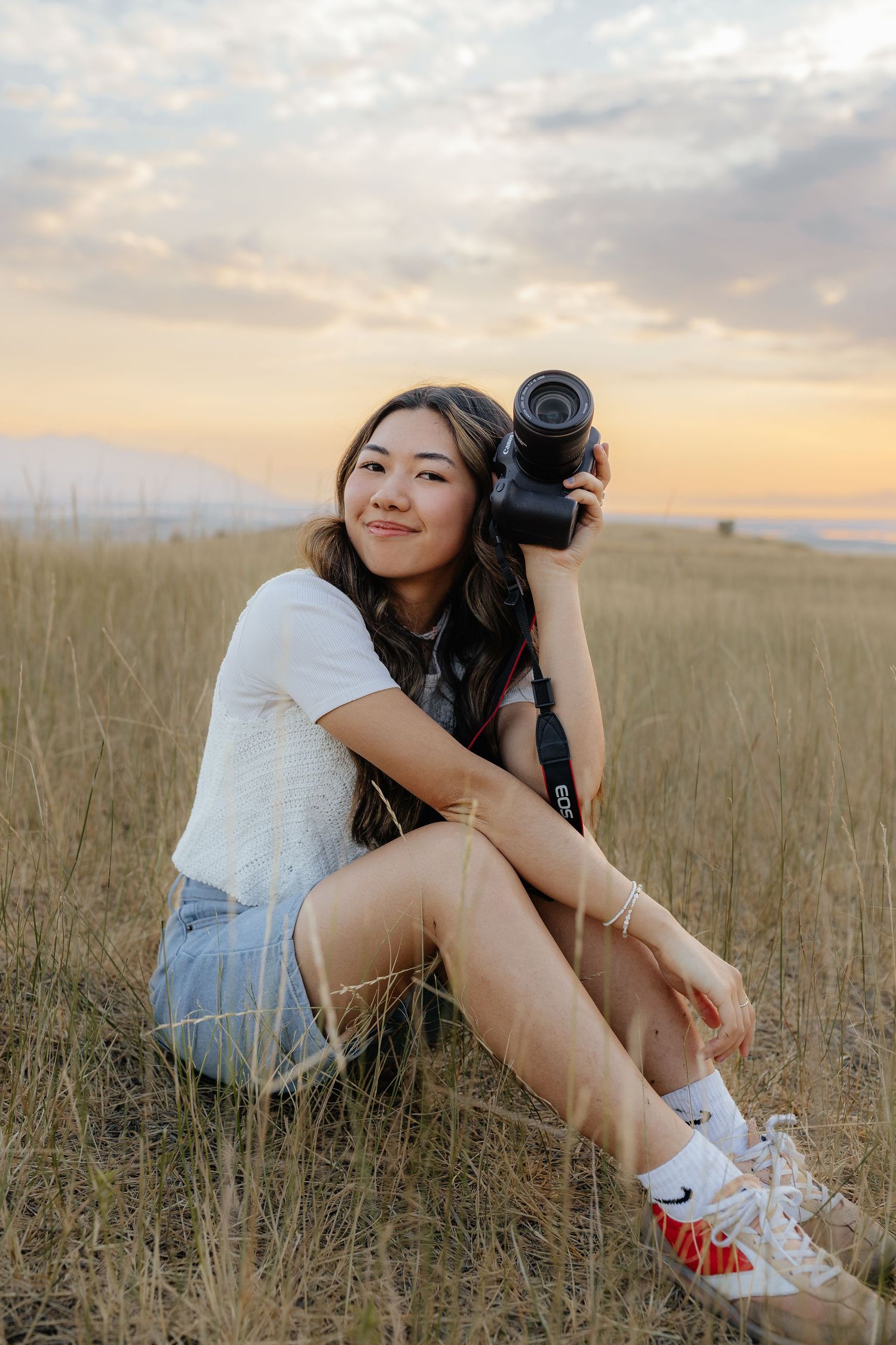Young woman sitting in a golden grass field at sunset holding a DSLR camera and smiling warmly.