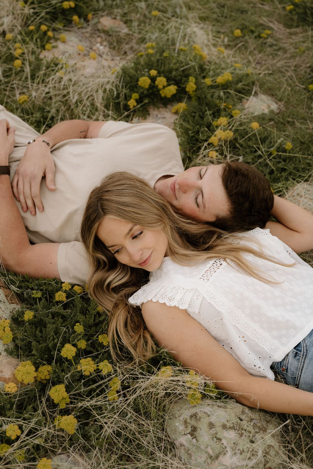 A couple lying in a meadow of yellow wildflowers, wearing casual white clothing and resting peacefully in nature.