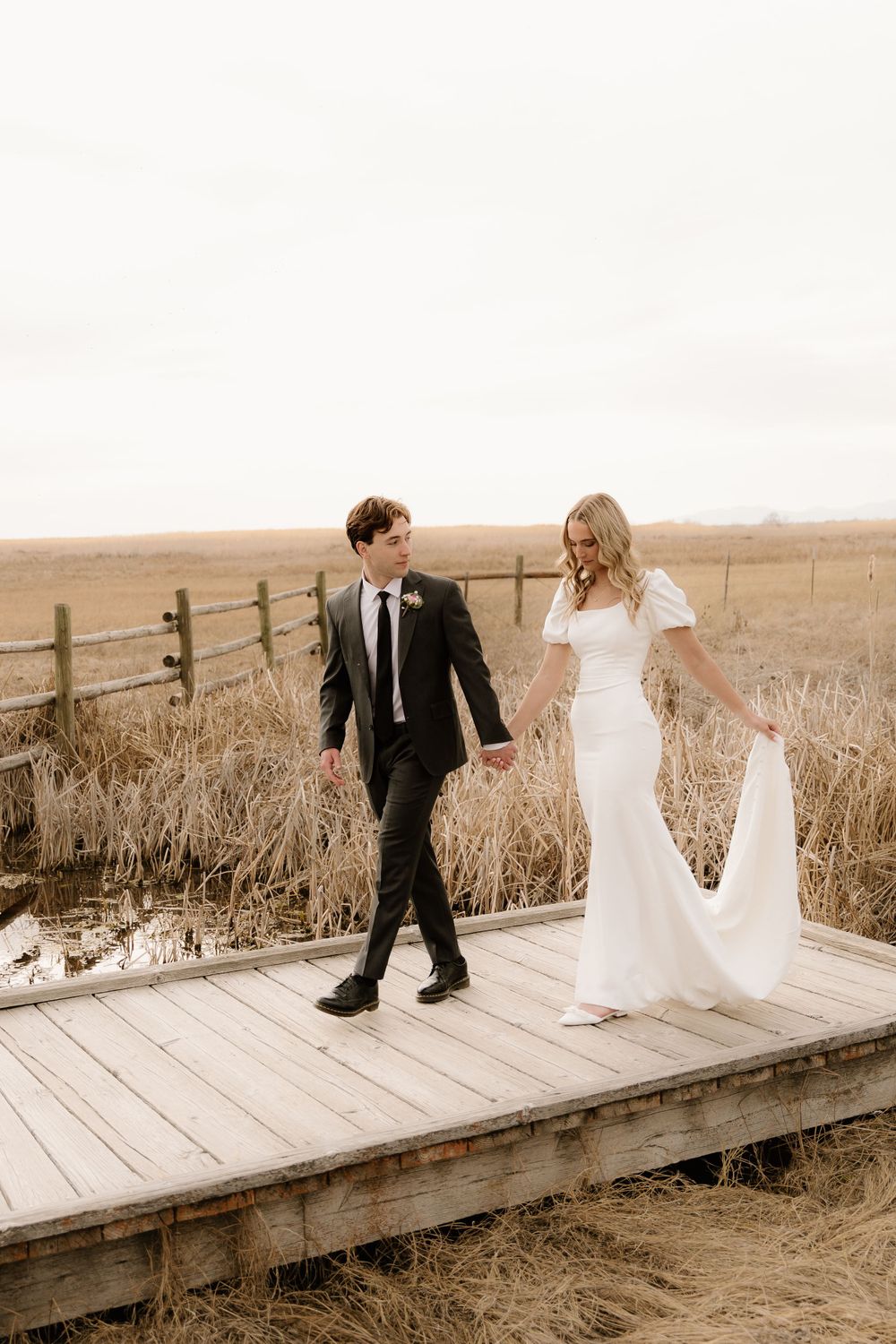 A couple in wedding attire walks hand in hand on a rustic wooden boardwalk surrounded by golden fields at sunset.