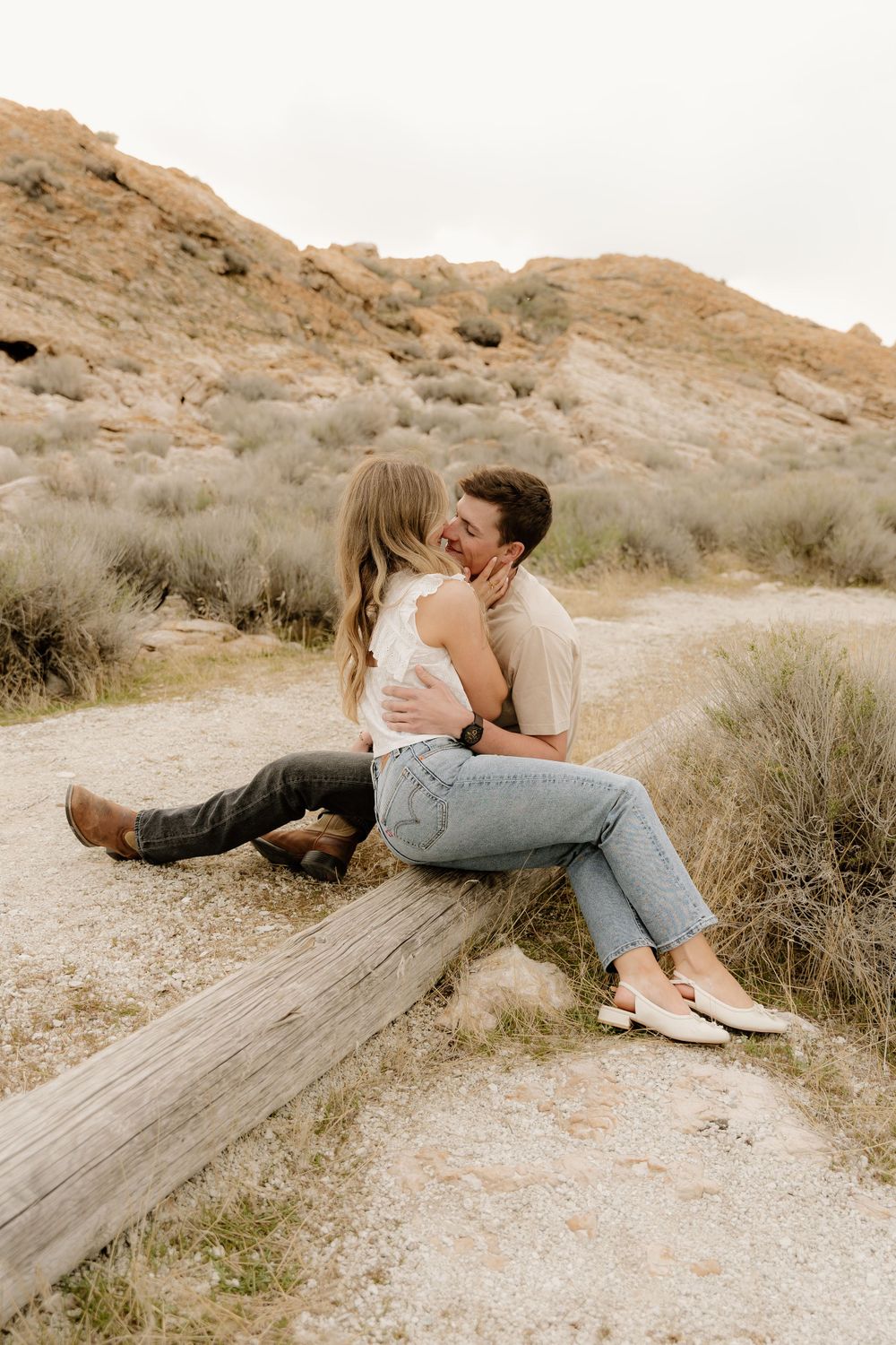 A romantic couple sharing an intimate moment while sitting on a fallen log in a desert landscape with rocky hills.