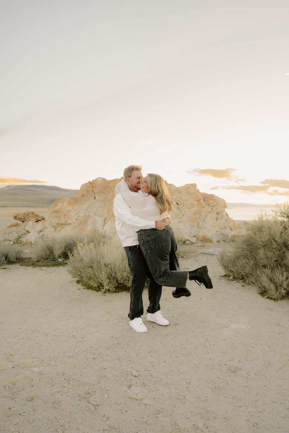Couple sharing a romantic kiss embrace in a desert landscape at golden hour with rocky formations and shrubs.