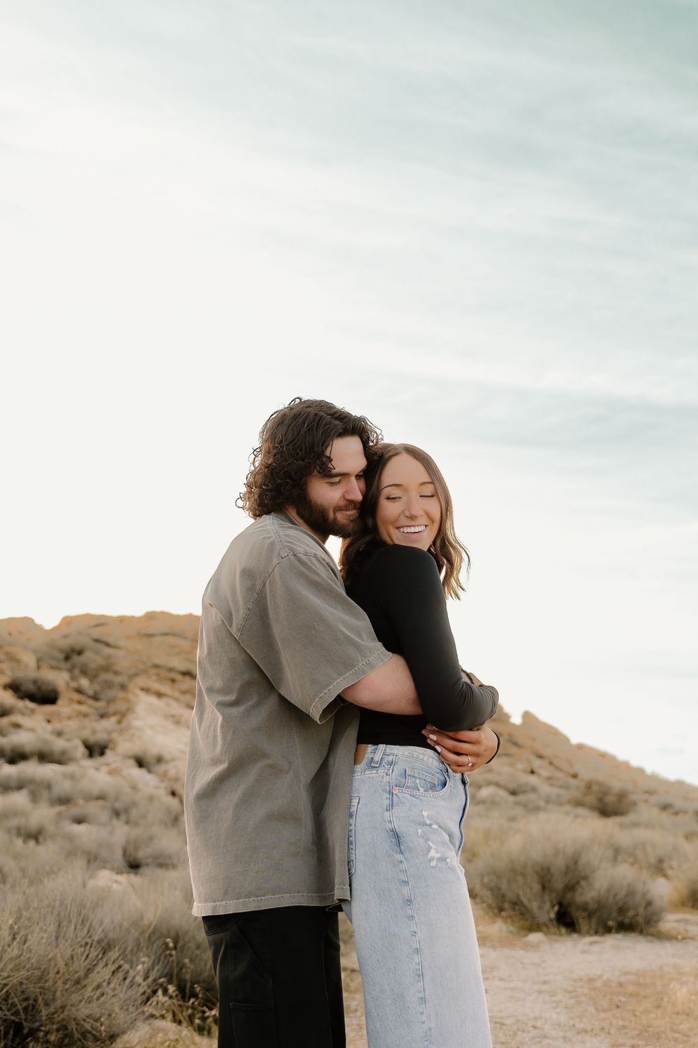 A couple embraces affectionately in a desert landscape with rocky hills and brush against an overcast sky.