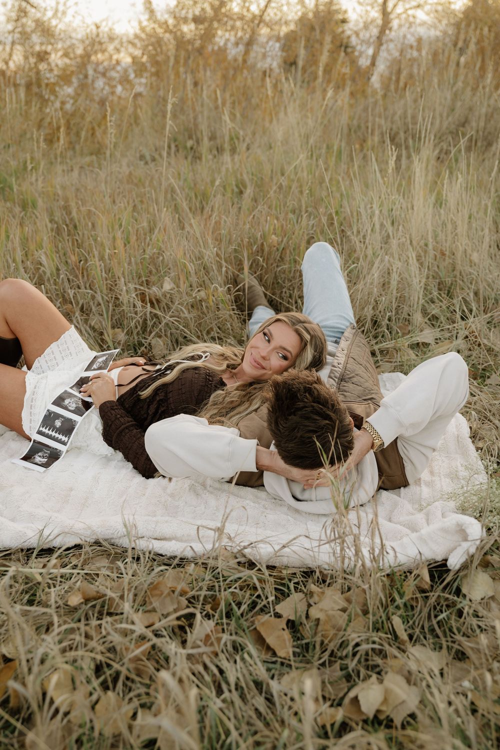 Couple lying on a white blanket in a field holding ultrasound photos, celebrating pregnancy outdoors.