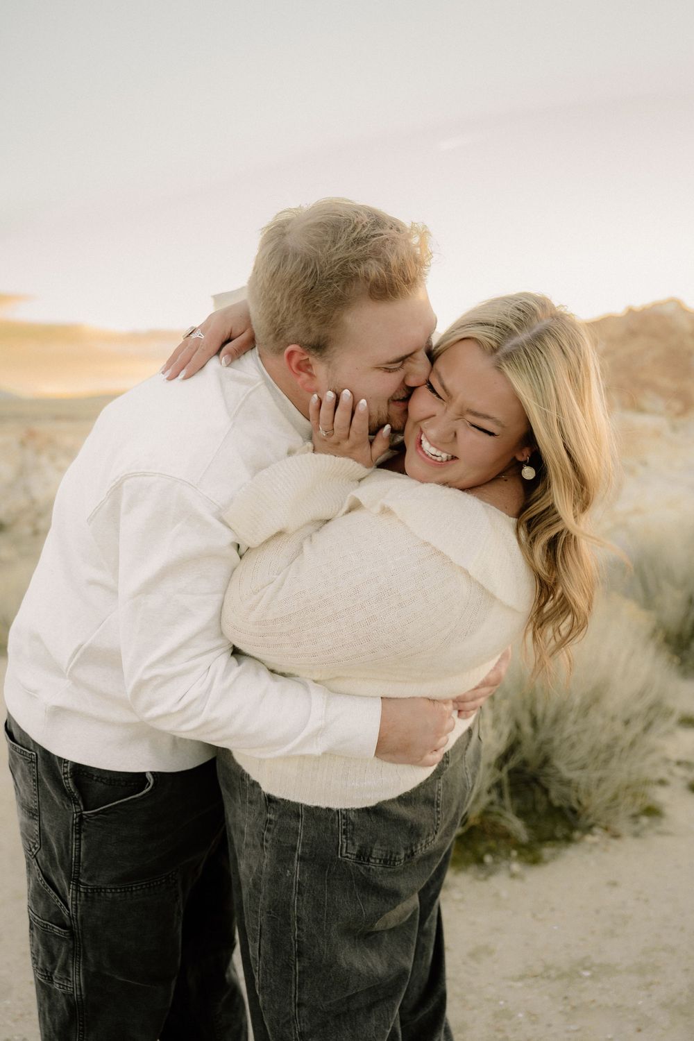 Couple embracing and laughing on sandy dunes at golden hour, woman in cream sweater and man in white shirt.
