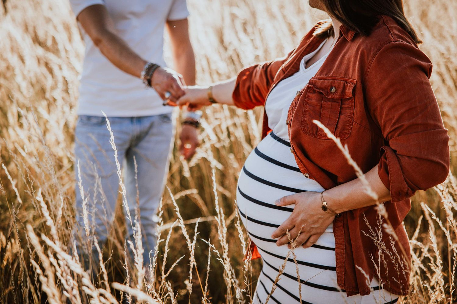 A couple holds hands while walking through a golden wheat field at sunset in casual autumn attire.