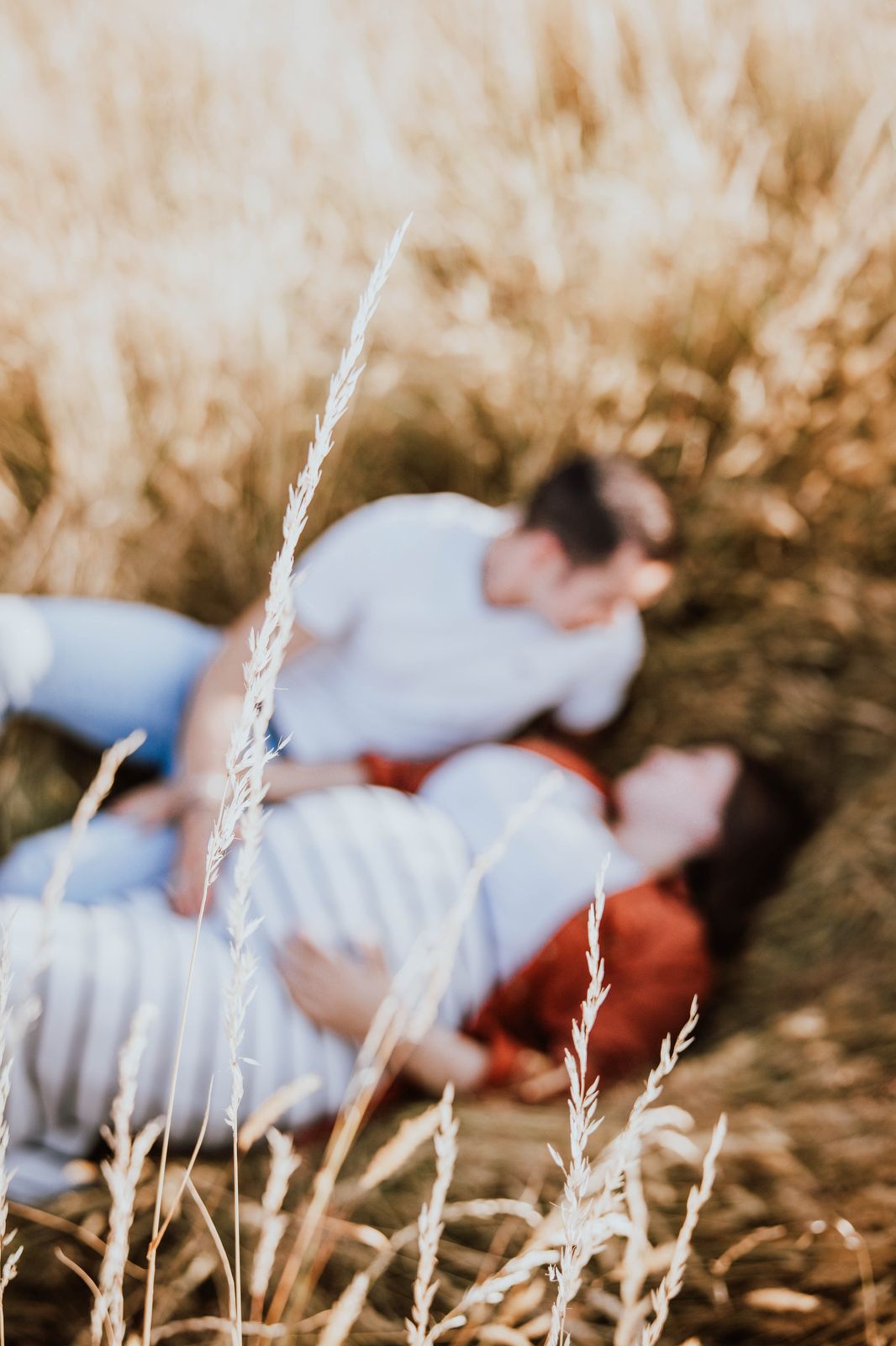 A couple lying in a field with tall white grass in the foreground creating a dreamy romantic atmosphere.