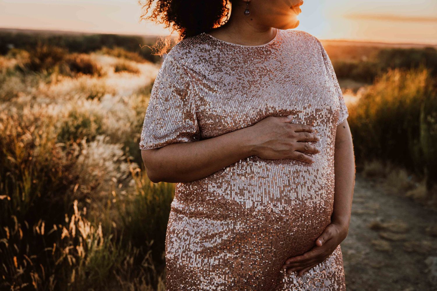Pregnant woman in sparkly rose gold dress cradles baby bump during golden hour sunset photoshoot in nature.