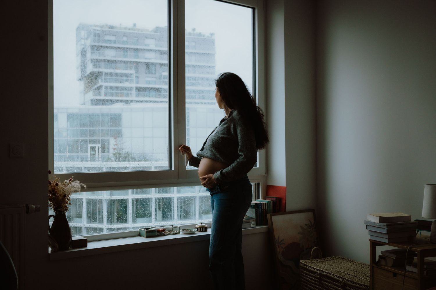 A person stands silhouetted against a window in a dimly lit room with a city view in the background.