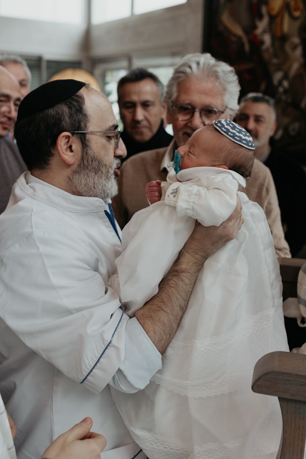 A Jewish bris ceremony shows a mohel in white holding a baby wrapped in white cloth during the traditional ritual.