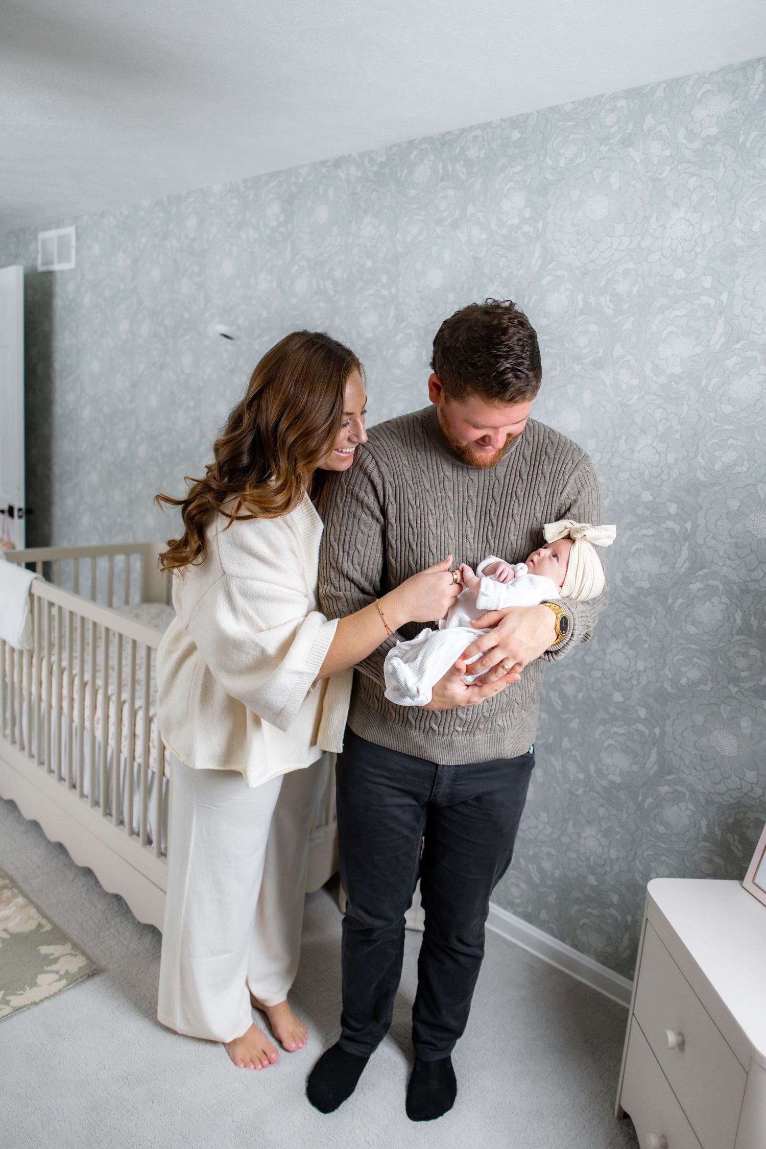 Parents in grey and white sweaters lovingly gaze at their newborn baby in a bright nursery setting.