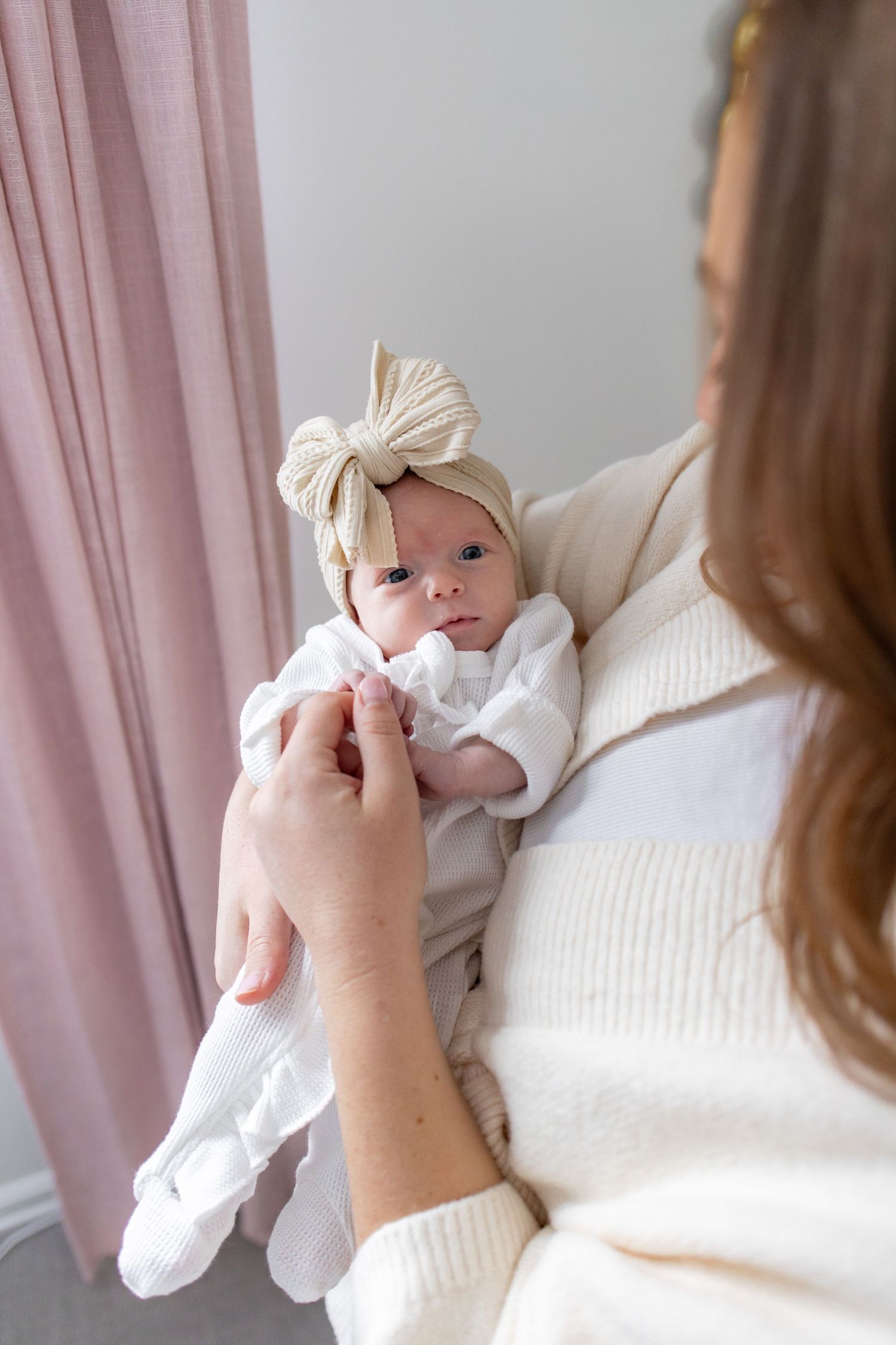 A baby wearing a white bow headband and white outfit is held by someone in a cream sweater near pink curtains.