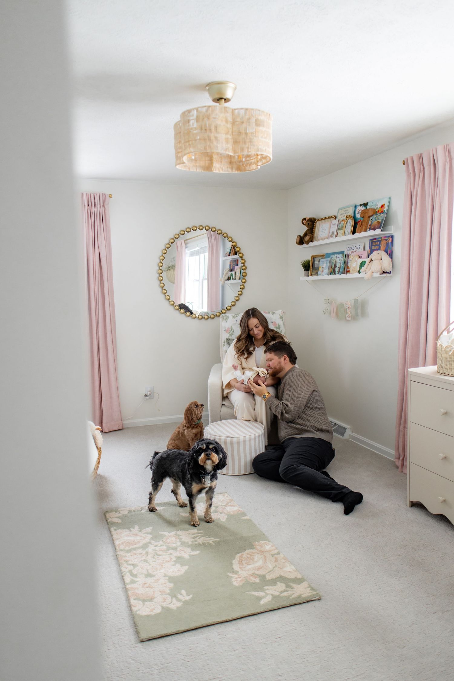 A cozy nursery room with pink curtains, decorative mirror, floating shelves, and two dogs sitting near a floral rug.