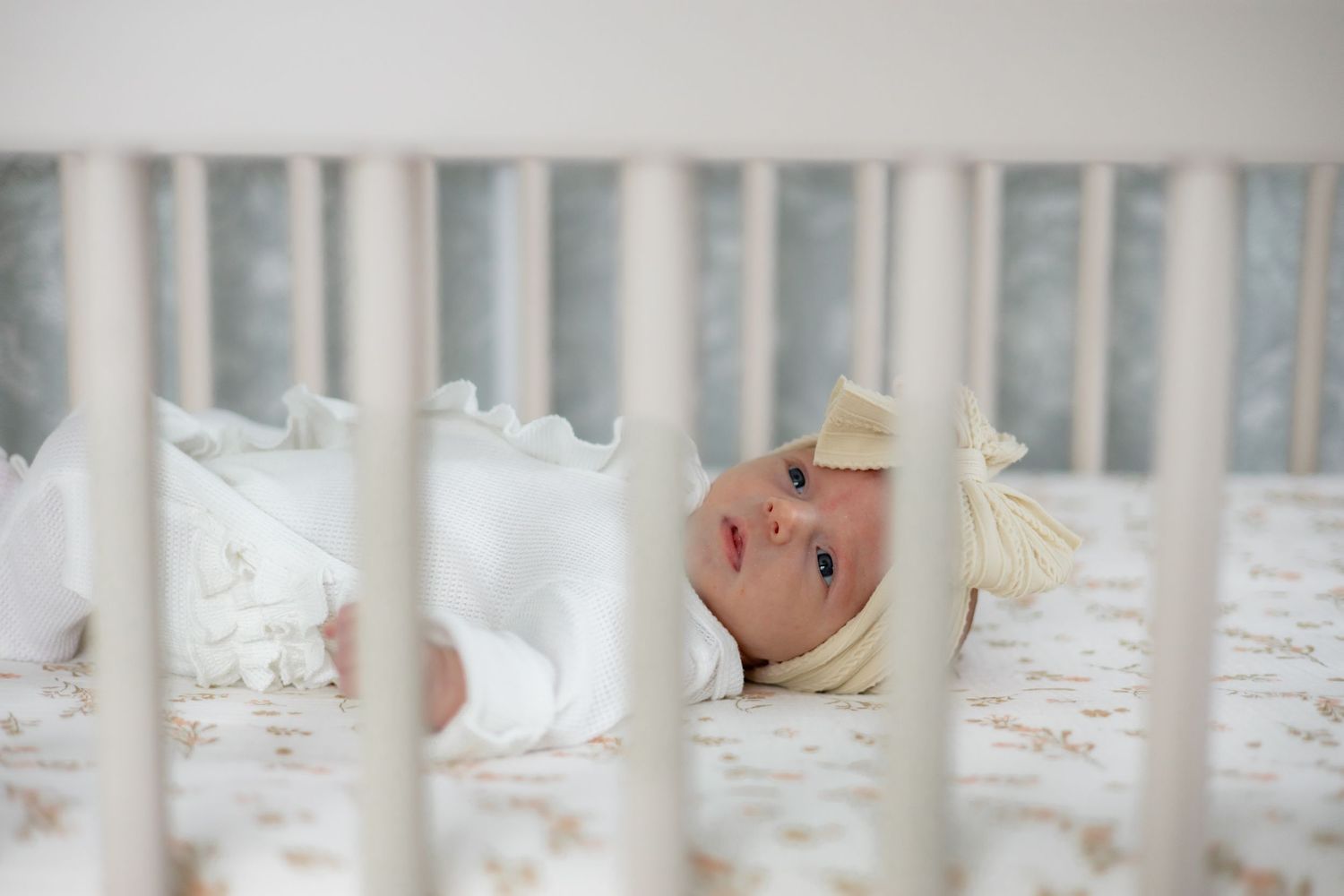 A sleeping baby wearing white clothing lies peacefully in a white crib with vertical slats.
