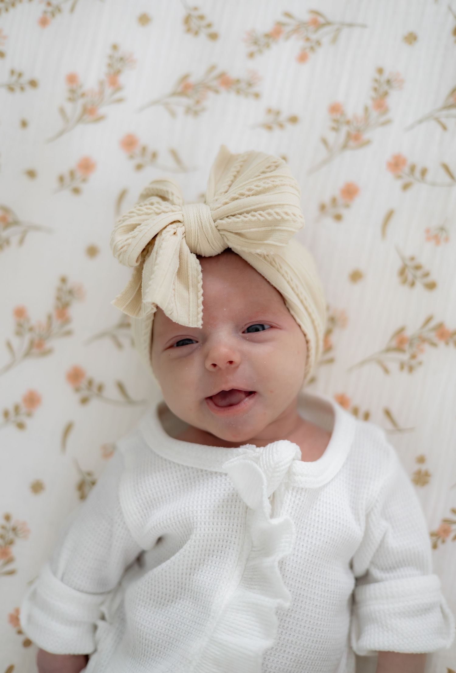 Baby wearing white outfit and cream bow headband lies on floral patterned fabric.