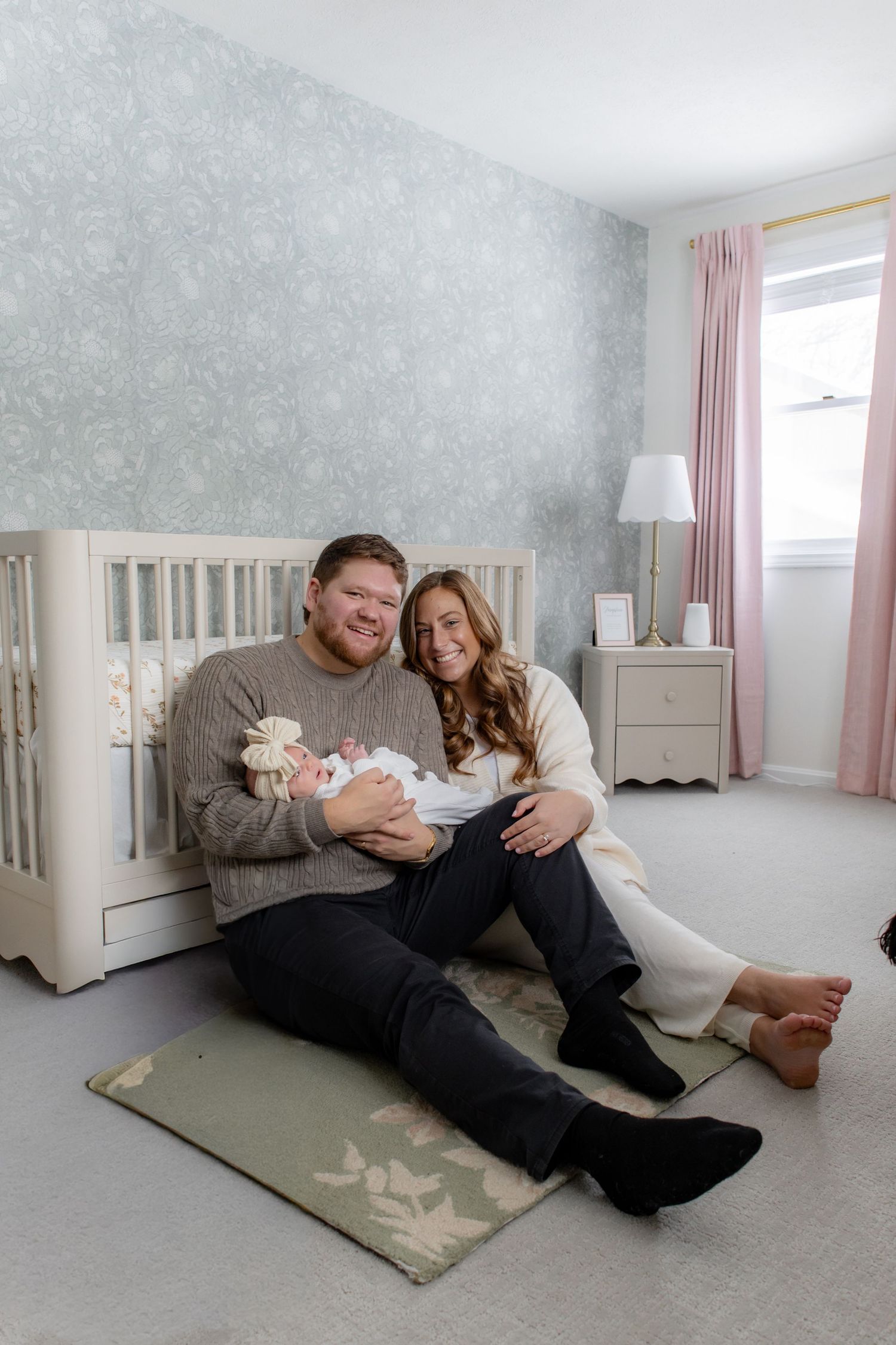 A nursery room decorated in soft gray and pink tones with a white crib and two people sitting on the floor together.