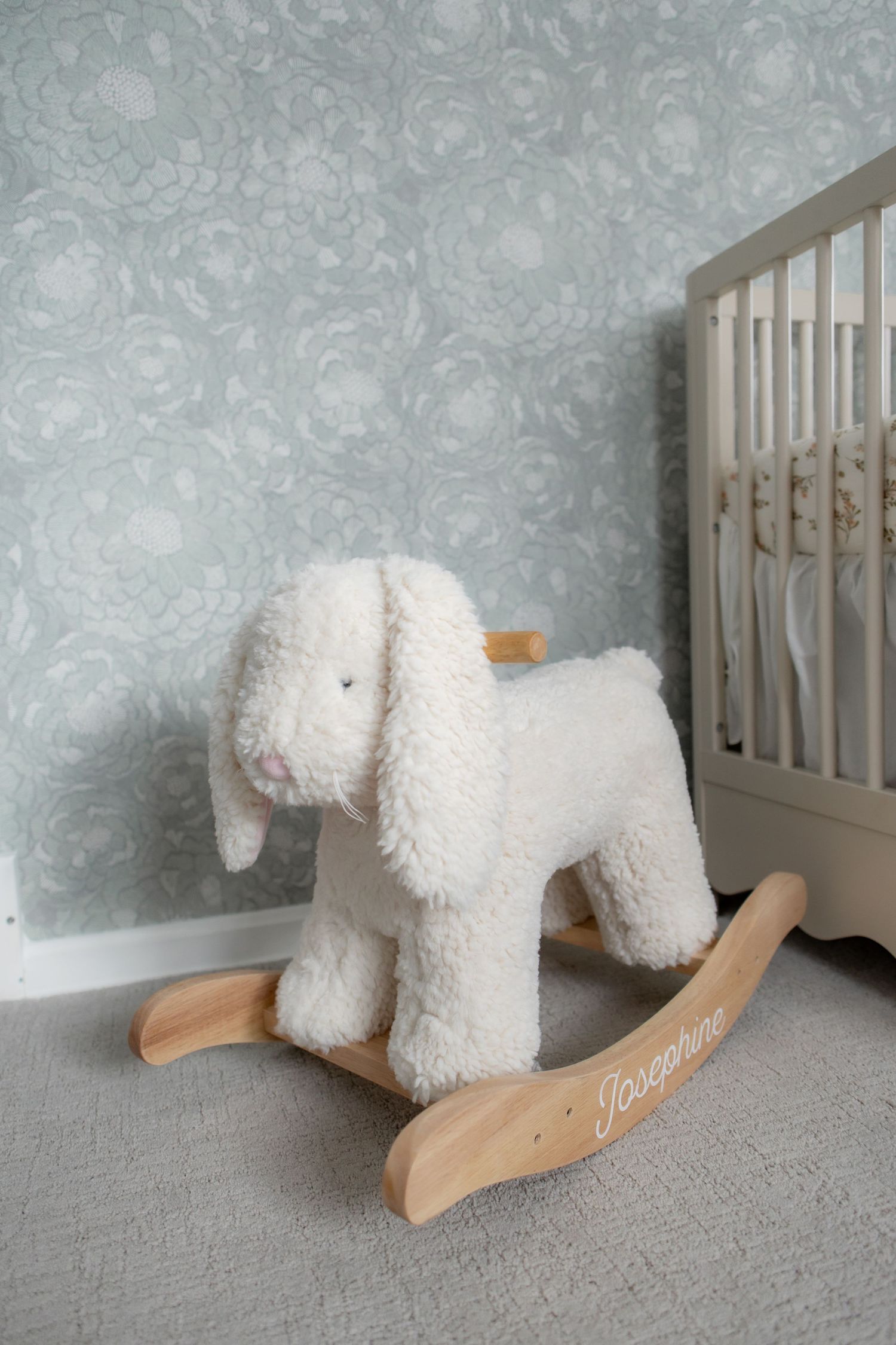A white plush rocking horse toy sits on gray carpet against a textured blue-gray wall in a nursery setting.